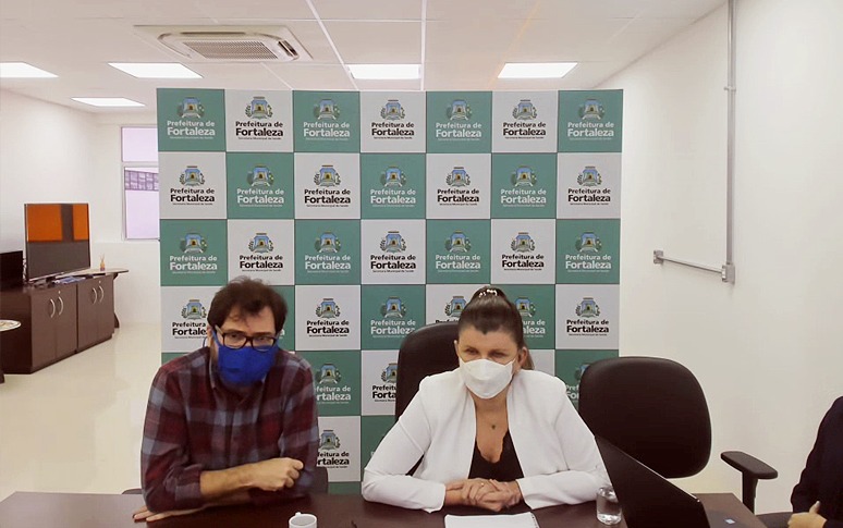 Antônio Lima Neto e Joana Maciel sentados lado a lado usando máscaras e backdrop da Prefeitura ao fundo